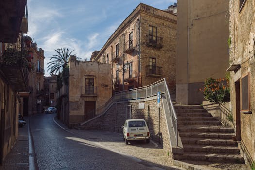 Sunlit street view in Caccamo, Sicily with classic architecture and cars.