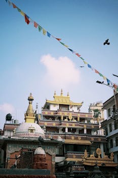 Vibrant prayer flags flutter over traditional Nepalese temple with ornate architecture under blue sky.